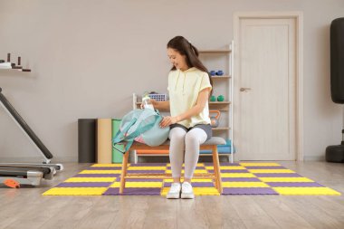 Sportif, genç ve mutlu Asyalı bir kadın, bir şişe protein içeceği ve poşeti ile spor salonunda bankta oturuyor.