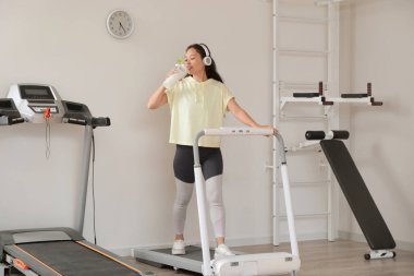 Spor salonundaki spor aletleri ve bir şişe protein içeceğiyle kulaklıklı mutlu bir Asyalı kadın.