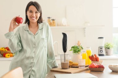 Elma ve el blendırlı genç bir kadın mutfakta kahvaltı için smoothie hazırlıyor.