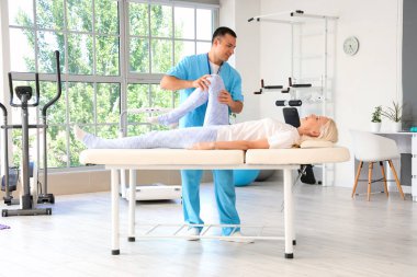 Erkek fizyoterapist rehabilitasyon merkezindeki kanepede olgun bir kadınla çalışıyor.