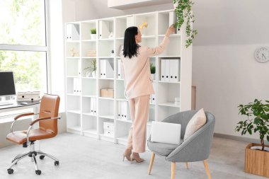 Genç bir kadın hafif ofisteki raflardan klasör alıyor.