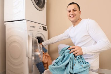 Çamaşır odasındaki çamaşır makinesine kirli çamaşır koyan genç adam.