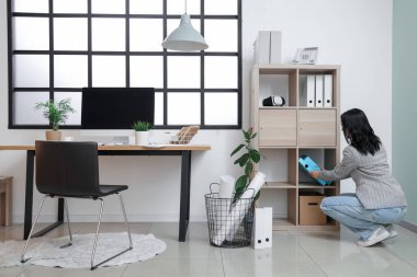 İş yerinin yakınındaki raflardan klasör alan genç bir kadın modern bilgisayarla güzel bir ofiste.