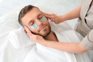Göz altı bantlı yakışıklı genç adam güzellik salonunda yüz masajı yaptırıyor.
