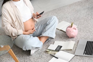 Cep telefonu ve kumbaralı hamile bir kadın evde yerde oturuyor, yakın plan. Annelik Faydası konsepti