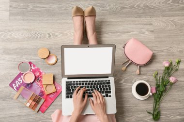 Makyaj sanatçısı kozmetik ürünleriyle ahşap zeminde dizüstü bilgisayar kullanıyor.