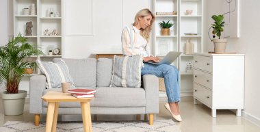 Orta yaşlı kadın evde kanepeüzerinde dizüstü bilgisayar kullanıyor