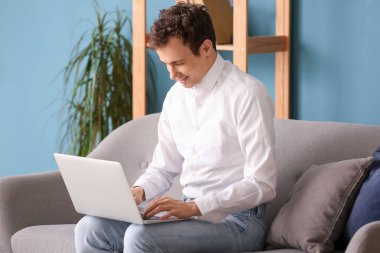 Evdeki kanepede oturan dizüstü bilgisayarı olan genç adam.