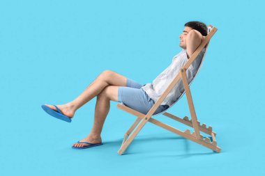 Genç adam mavi arka planda güverte sandalyesinde dinleniyor.