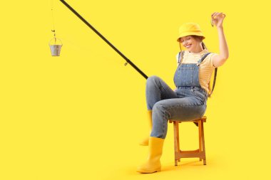 Happy young woman with fishing rod and small bucket sitting on stool against yellow background clipart