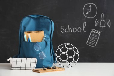 Siyah karatahtanın yanındaki beyaz masada kırtasiye malzemesi ve moleküler modelli mavi okul çantası.