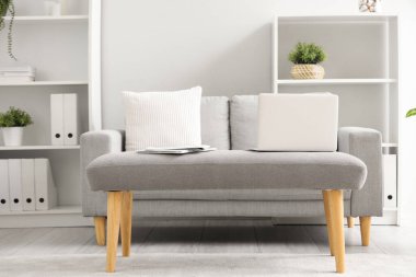 Laptop with newspapers on soft bench in light living room