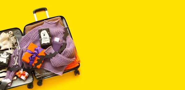 stock image Open suitcase with clothes, different Halloween decorations and gifts on yellow background with space for text