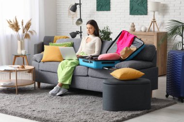 Evdeki koltukta bavulun yanında dizüstü bilgisayar kullanan genç bir kadın. Yaz yolculuğu konsepti