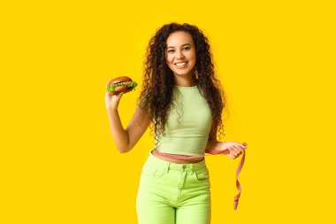 Burger yiyen ve sarı arka planda mezurası olan mutlu Afro-Amerikan kadın. Diyet kavramı