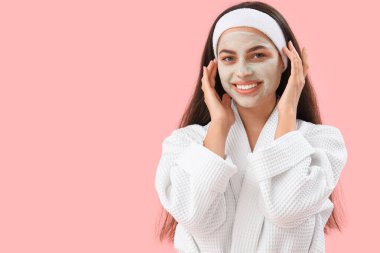 Pembe arka planda uygulamalı kil maskeli bornozlu güzel, genç ve mutlu bir kadın.