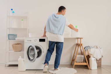 Genç adam çamaşır odasındaki masadan bir şişe deterjan alıyor.