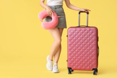 Sarı arka planda bavulu ve boyun yastığı olan genç bir kadın.