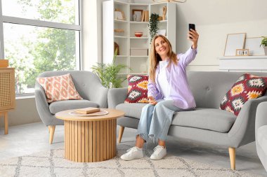 Cep telefonlu olgun bir kadın evdeki koltukta selfie çekiyor.