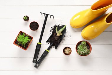 Beyaz ahşap arka planda tırmık, kürek ve lastik botlu bahçe aletleri. Üst görünüm