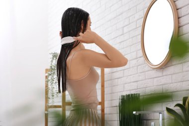 Güzel genç bir kadın banyoda aynanın yanında saçını maskeyle tarıyor.