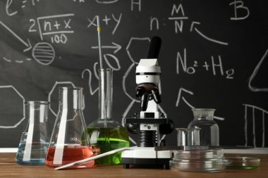 Modern mikroskop, petri kapları ve mataralar kimyasal formüllü karatahtanın yanındaki ahşap masada.