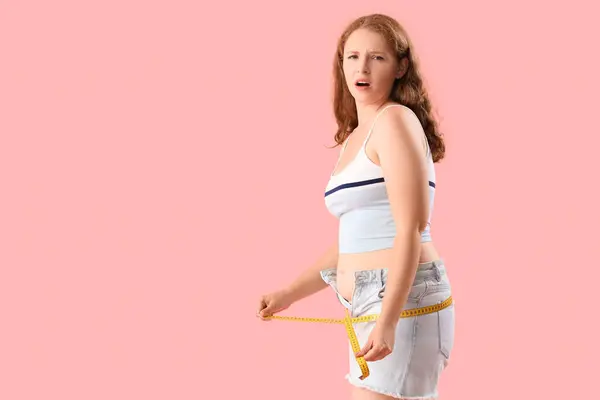 stock image Upset young woman in tight shorts measuring her hips on pink background. Weight gain concept