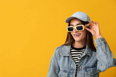 Şık güneş gözlüklü ve sarı arka planda şapkalı mutlu genç kadın.