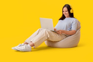 Kulaklıklı genç bir kadın, sarı arka planda, puf çantasına dizüstü bilgisayar kullanıyor.
