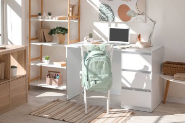 Kırtasiyeli, sırt çantalı ve laptoplu modern okul masası beyaz duvarın yanında.