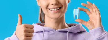 Young woman with plastic tooth showing thumb-up on blue background, closeup