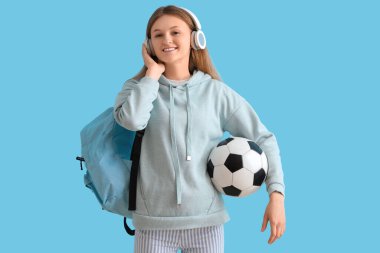 Sırt çantalı ve futbol toplu bir kız öğrenci mavi arka planda kulaklıkla müzik dinliyor.