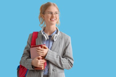 Sırt çantası, kitapları ve kulaklıkları mavi arka planda olan mutlu kız öğrenci.