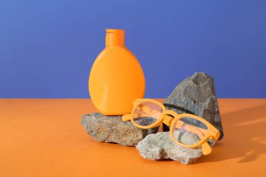 Bir şişe güneş kremi, güneş gözlüğü ve taşlarla mavi duvarın yanındaki turuncu masada.