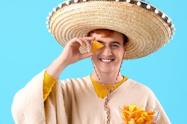 Sombrero giymiş genç Meksikalı bir adamın portresi ve mavi arka planda nachos.