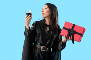 Cadı gibi giyinmiş genç bir Asyalı kadın ve mavi arka planda Cadılar Bayramı hediyesi.