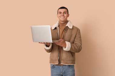 Bej arka planda dizüstü bilgisayarı olan koyun derisi ceketli genç bir adam.