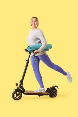 Güzel, olgun, sportif bir kadın. Sarı arka planda scooter ve yoga minderi var.