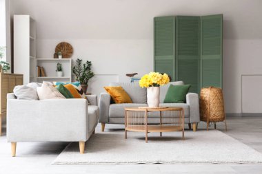 Aydınlık oturma odasının içinde gri kanepeler ve ahşap sehpanın üzerinde sarı narsisli çiçekler.