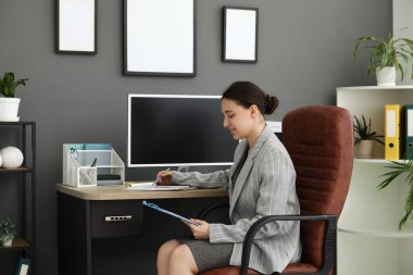 Elinde pano olan kadın ofis çalışanı koltukta oturuyor ve şık bir ofiste evrak işleriyle uğraşıyor.