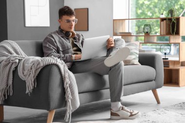 Gözlüklü genç adam evdeki kanepede dizüstü bilgisayar kullanıyor.