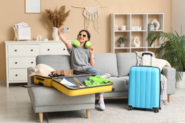 Güneş gözlüklü genç bir kadın cep telefonuyla selfie çekiyor ve evdeki kanepede bavul hazırlıyor. Yaz yolculuğu konsepti