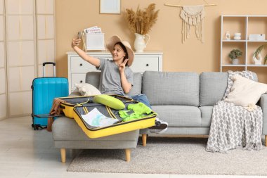 Şapkalı genç bir kadın cep telefonuyla selfie çekiyor evdeki kanepede bavulun yanında. Yaz yolculuğu konsepti