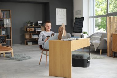 Genç erkek programcı cep telefonu ve bir fincan kahveyle modern ofis işyerinde dinleniyor.