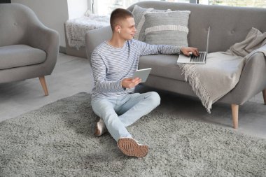 Tablet bilgisayarı ve dizüstü bilgisayarı olan genç bir erkek serbest yazar modern ofis ofisindeki kanepenin yanında oturuyor.