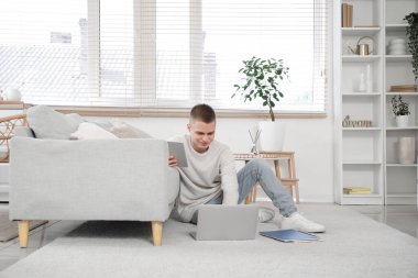 Tablet bilgisayarı ve dizüstü bilgisayarı olan genç bir erkek ev ofisteki kanepenin yanında oturuyor.