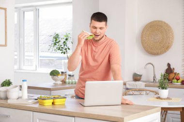 Laptoplu ve beslenme çantalı genç adam mutfakta lezzetli sandviçler yiyor.