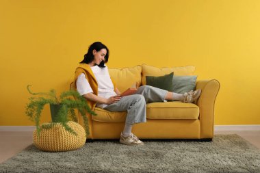 Evdeki sarı kanepede kitap okuyan güzel genç bir kadın.