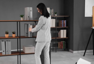 Genç bir kadın şık ofisteki raflardan klasör alıyor.