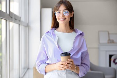 Gözlüklü genç bir kadın. Evinin penceresinin yanında bir fincan kahve.